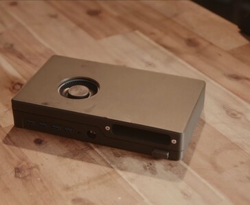 Una console di gioco nera e compatta, dotata di lettore disco integrato, porte USB e ventola di raffreddamento posizionata sulla parte superiore, appoggiata su un tavolo di legno.
