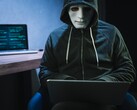 Una figura mascherata e incappucciata lavora su un computer portatile in una stanza buia.