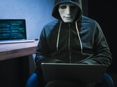 Una figura mascherata e incappucciata lavora su un computer portatile in una stanza buia.