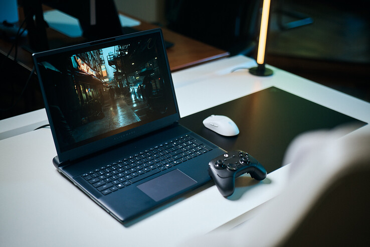 Un laptop da gaming Alienware aperto, posizionato su una scrivania bianca da studio, su cui viene riprodotta una scena di strada scura e riflettente con dettagli nitidi ottenuti tramite path tracing, abbinato a un controller nero e a un mouse wireless bianco.