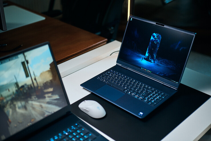 Un portatile da gaming di fascia media di colore blu scuro (Alienware Aurora 16X) inclinato su un tappetino da scrivania con uno sfondo blu luminoso in stile fantascientifico, posizionato accanto a un mouse wireless bianco con un altro portatile sfocato in primo piano.
