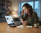 Una donna seduta a una scrivania guarda il proprio smartphone; accanto ad esso ci sono un computer portatile, alcune ricevute e un blocco note
