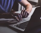 Mani che digitano sulla tastiera di un MacBook in condizioni di scarsa illuminazione, con il logo Apple che risplende