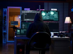 Un ricercatore incappucciato lavora su più schermi in un ambiente buio. Nella foto un hacker generico.