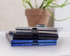 Pila di smartphone pieghevoli che rappresentano il concetto del prossimo Apple iPhone Fold, posizionati su un tavolo bianco con una pianta in vaso sullo sfondo (Fonte: Notebookcheck)