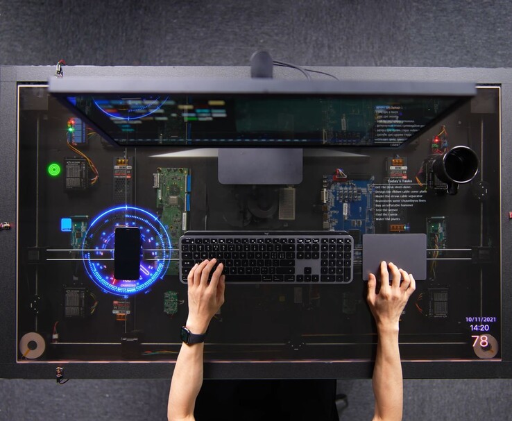 La scrivania fai-da-te di HTX Studio, ispirata ad AirPower, in uso, con uno smartphone in ricarica sul display trasparente accanto a una tastiera, un trackpad e un monitor.