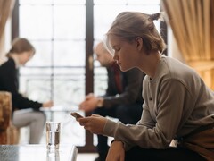 Un'adolescente guarda il suo smartphone, mentre sullo sfondo due adulti siedono fuori fuoco.