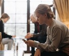 Un'adolescente guarda il suo smartphone, mentre sullo sfondo due adulti siedono fuori fuoco.