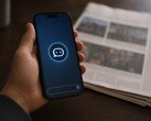 Una mano regge uno smartphone su cui è visibile l’icona di un chatbot; accanto ad esso giace un giornale cartaceo.
