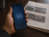 Una mano regge uno smartphone su cui è visibile l’icona di un chatbot; accanto ad esso giace un giornale cartaceo.