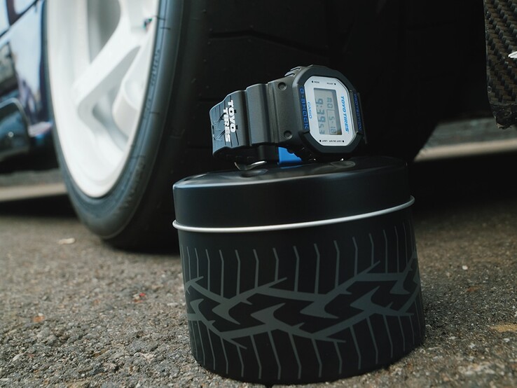 Orologio Casio G-Shock DW5600TT25-1 x Toyo Tires esposto su una scatola di metallo a tema pneumatici accanto a una ruota di automobile.