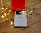 Un OnePlus 15 è appoggiato su un tavolo di legno, circondato da calde lucine a filo, con una confezione di vendita OnePlus di colore rosso vivo posizionata sullo sfondo.