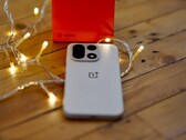Un OnePlus 15 è appoggiato su un tavolo di legno, circondato da calde lucine a filo, con una confezione di vendita OnePlus di colore rosso vivo posizionata sullo sfondo.