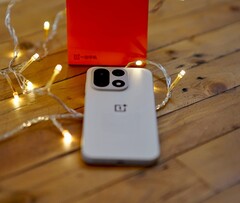 Un OnePlus 15 è appoggiato su un tavolo di legno, circondato da calde lucine a filo, con una confezione di vendita OnePlus di colore rosso vivo posizionata sullo sfondo.