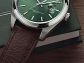 Primo piano dell'orologio Timex Legacy Day and Date, che mostra il quadrante verde soleil, la lunetta color argento e le anse spazzolate, la corona zigrinata e il cinturino in pelle marrone testurizzata, su uno sfondo con una scatola verde scuro e una superficie in legno.