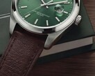 Primo piano dell'orologio Timex Legacy Day and Date, che mostra il quadrante verde soleil, la lunetta color argento e le anse spazzolate, la corona zigrinata e il cinturino in pelle marrone testurizzata, su uno sfondo con una scatola verde scuro e una superficie in legno.