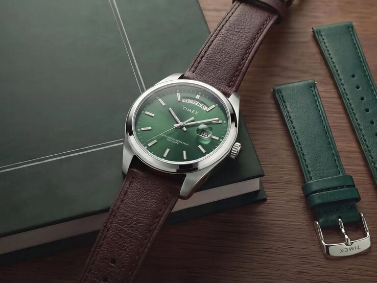 Il Timex Legacy Day and Date con quadrante verde e cinturino in pelle marrone esposto su un libro verde scuro accanto al cinturino intercambiabile in pelle verde.