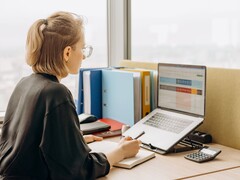 Una donna con gli occhiali lavora su un foglio di calcolo al computer portatile, con dei raccoglitori e una calcolatrice sulla scrivania