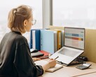 Una donna con gli occhiali lavora su un foglio di calcolo al computer portatile, con dei raccoglitori e una calcolatrice sulla scrivania