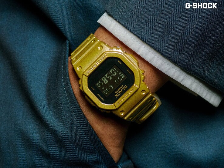 Primo piano di un orologio Casio G-Shock DW-5600BEP-9 dorato indossato al polso su uno sfondo di tessuto blu scuro. L’orologio presenta un display digitale nero, cassa e cinturino dorati e il marchio BlackEyePatch sulla lunetta e sul quadrante.