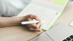 Una persona sta scrivendo appunti su un taccuino accanto a un computer portatile aperto