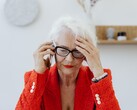 Una signora anziana dai capelli bianchi e con gli occhiali, al telefono in un luminoso salotto, con la mano appoggiata alla tempia.