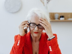 Una signora anziana dai capelli bianchi e con gli occhiali, al telefono in un luminoso salotto, con la mano appoggiata alla tempia.