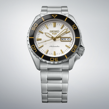 Seiko 5 Sports SKX HDB004 con quadrante bianco-argento, lunetta nera con indicazioni color oro, lancette e indici dorati e bracciale in acciaio inossidabile, mostrato su uno sfondo grigio chiaro.